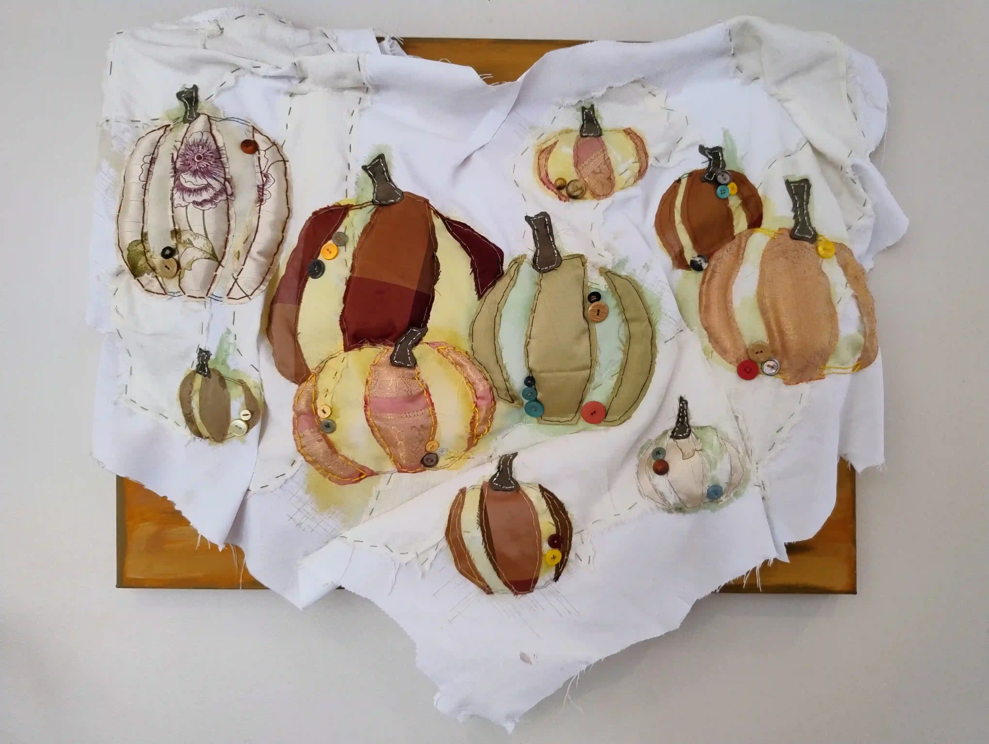 A white piece of cloth with various colours of pumpkins sewn onto it. The cloth is attatched to an orange canvas